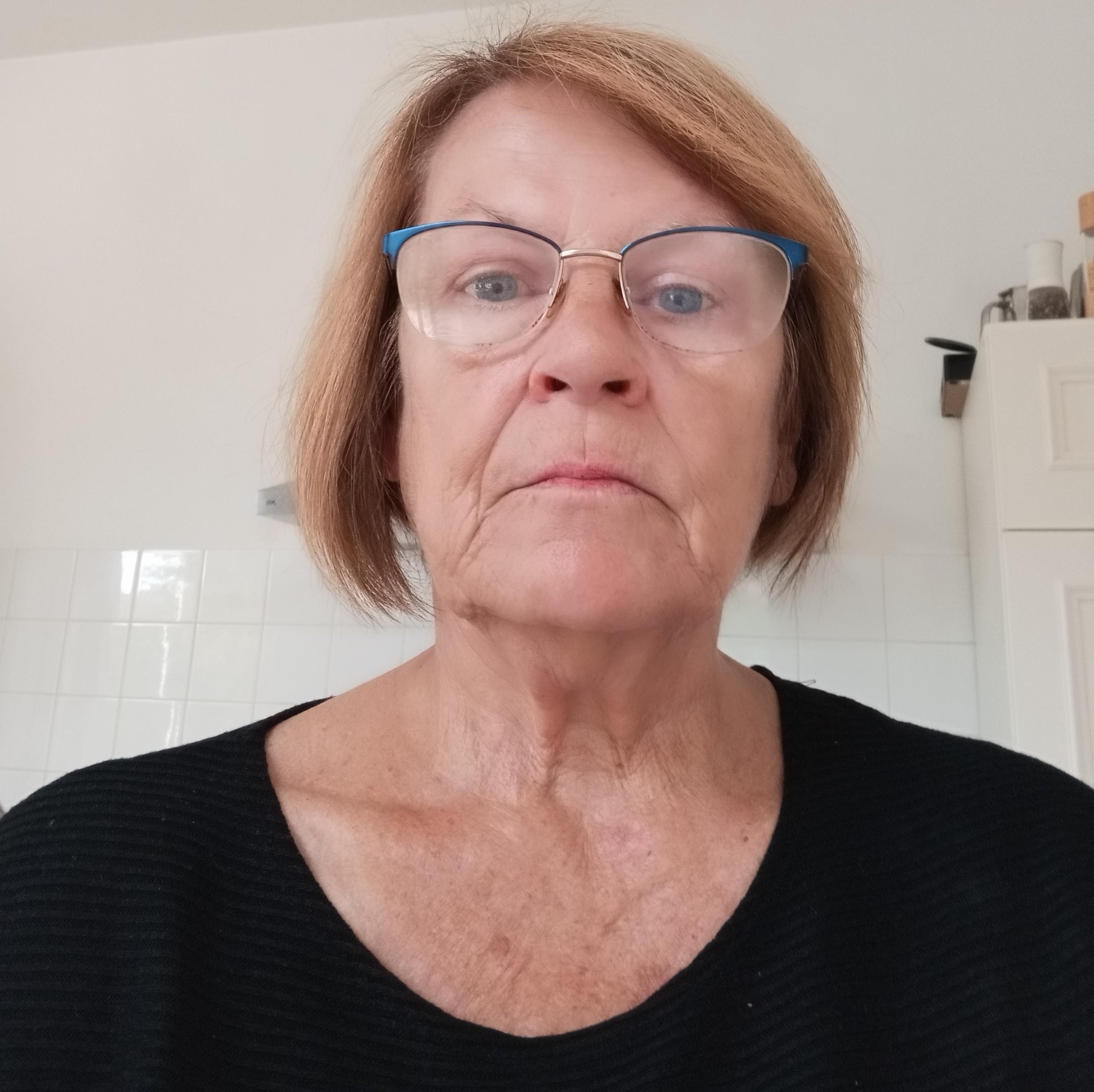 Suus draagt een zwart shirt een een bril met een half blauw montuur. Ze heeft half lang haar. Op de achtergrond is een witte tegelmuur te zien.