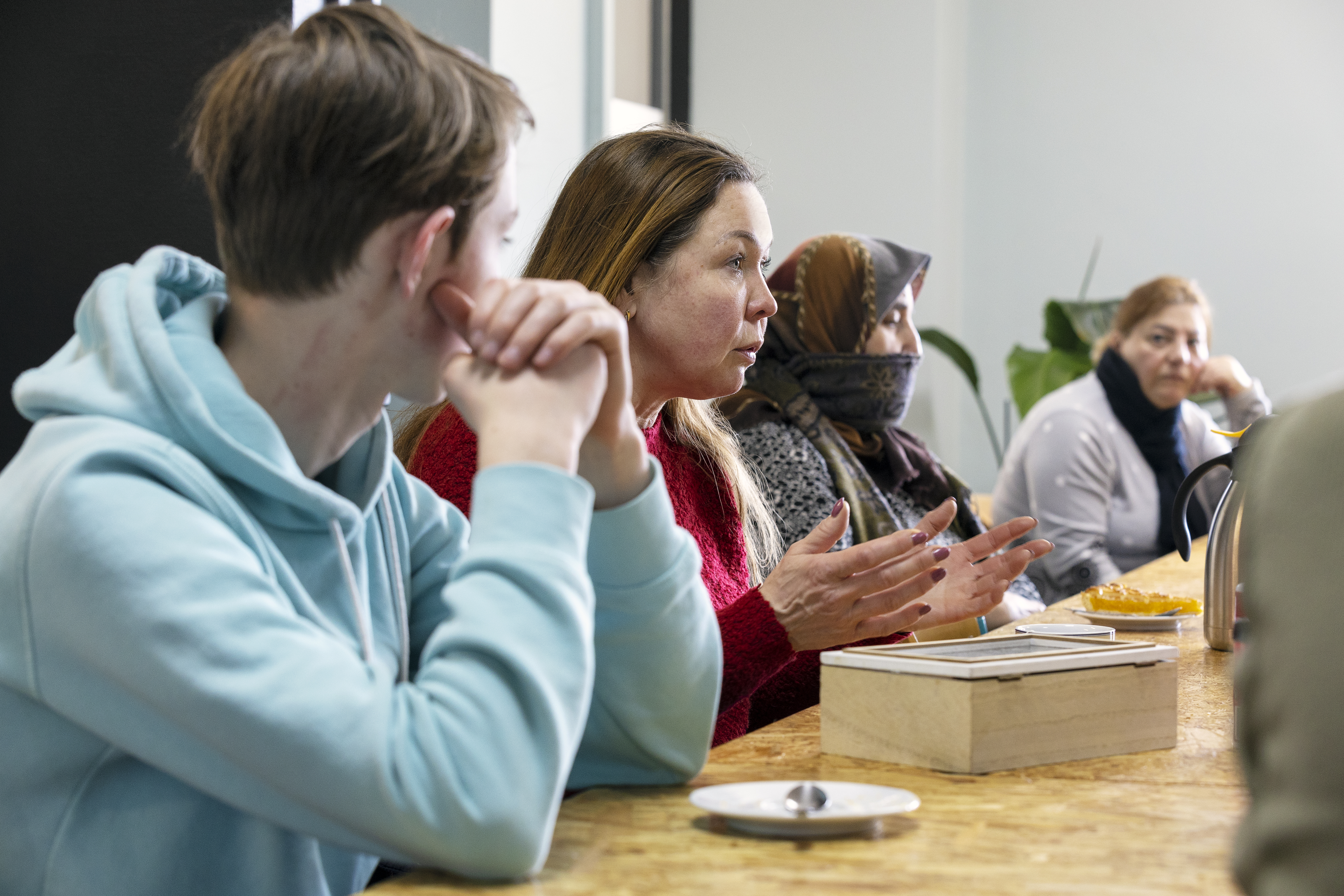 Een vrouw aan een tafel legt iets uit. Op de voor- en achtergrond zijn meer mensen die aan tafel zitten te zien.
