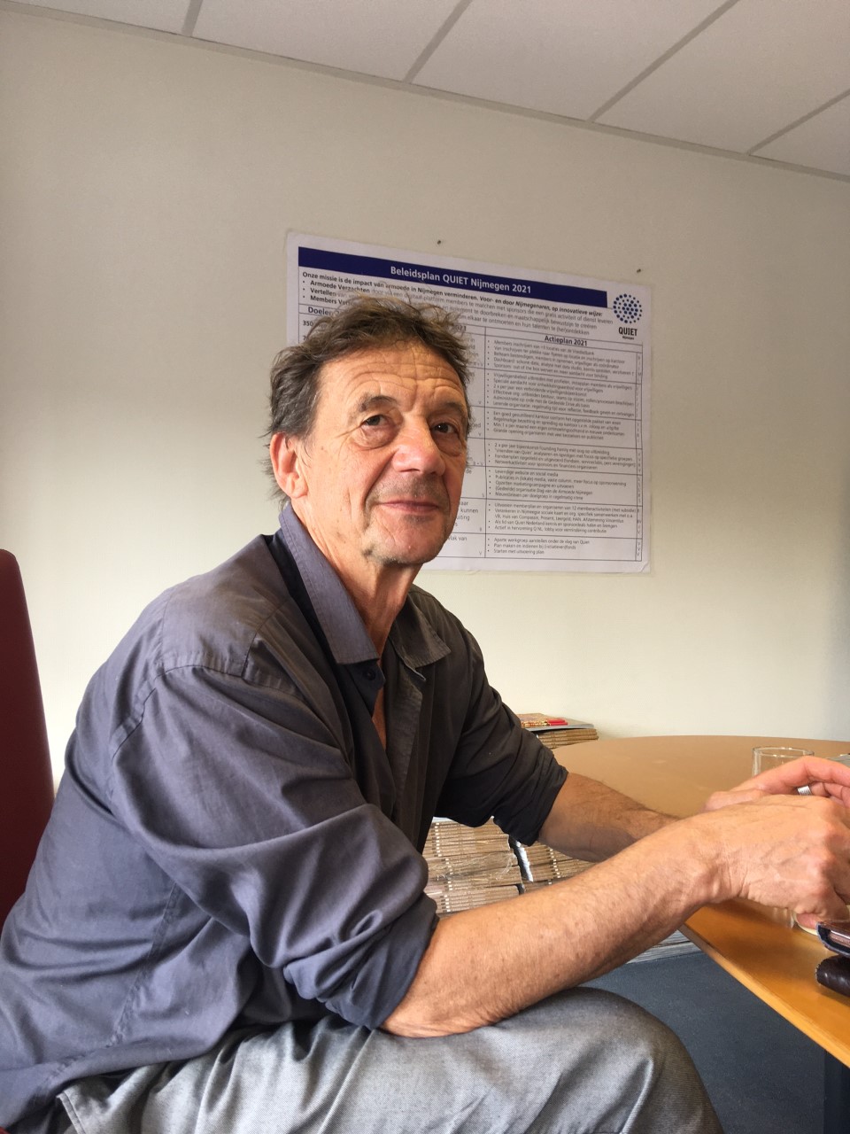 Henks handen liggen op de tafel voor hem. Hij kijkt sympathiek in de camera. Henk draagt een blauwe blouse en een grijze broek.