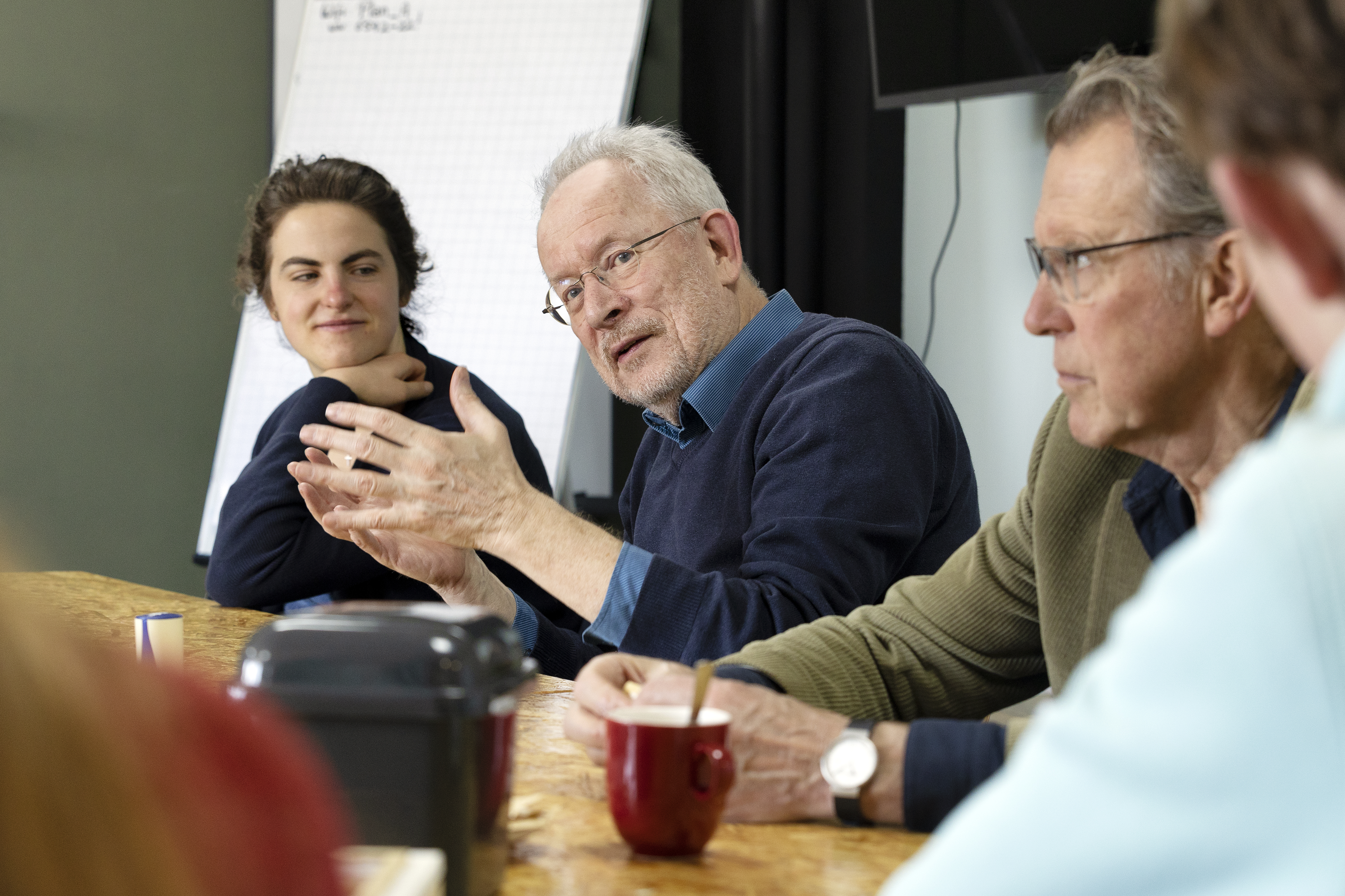 Huisarts Guus legt iets uit. Hij maakt handgebaren. Rondom hem zitten meer mensen aan tafel naar hem te luisteren.