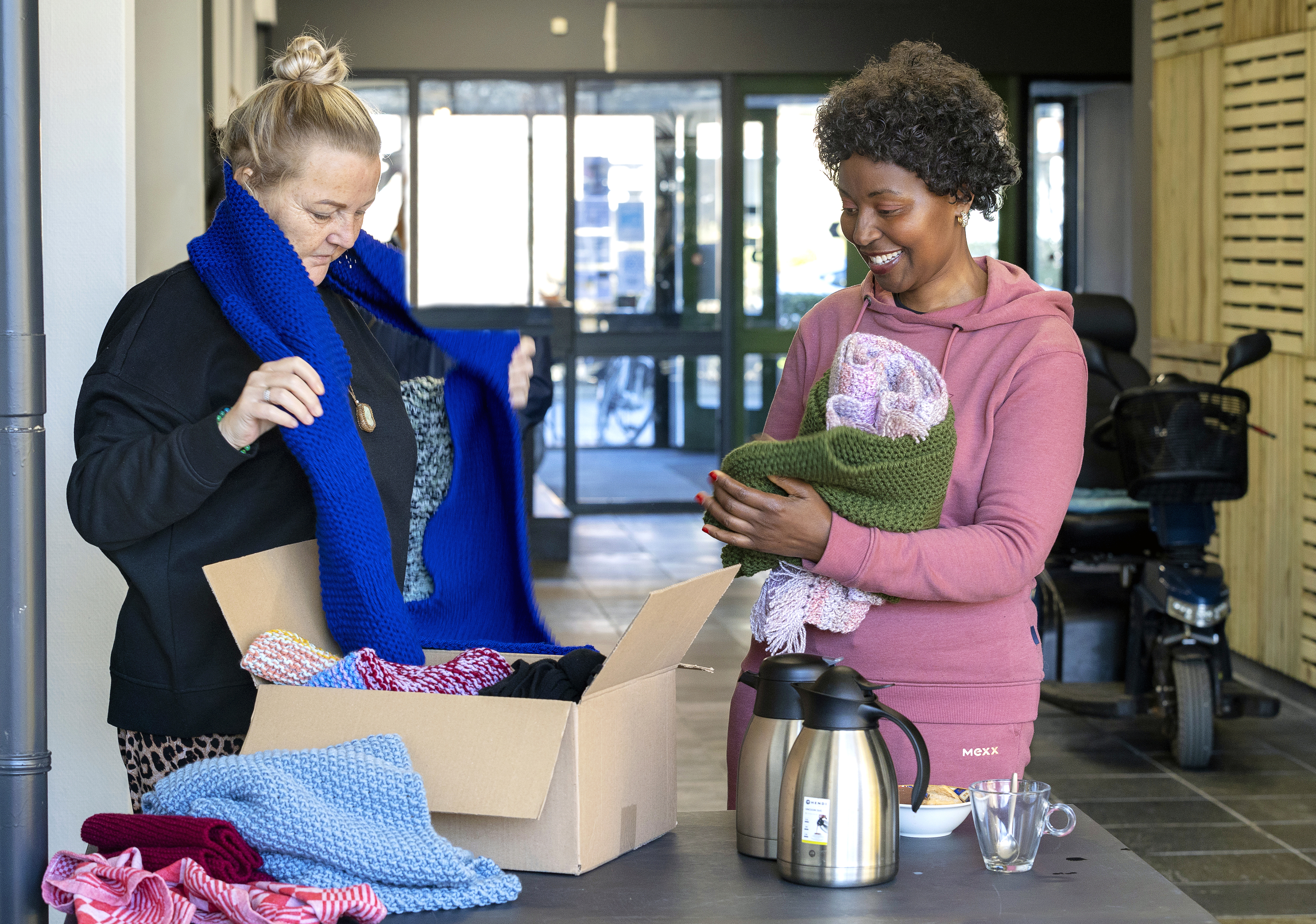 twee vrouwen halen gebreide sjaals uit een doos. Ze proberen ze om. Op de achtergrond zijn een scootmobiel en de gang van het kantorenpand van Quiet te zien.
