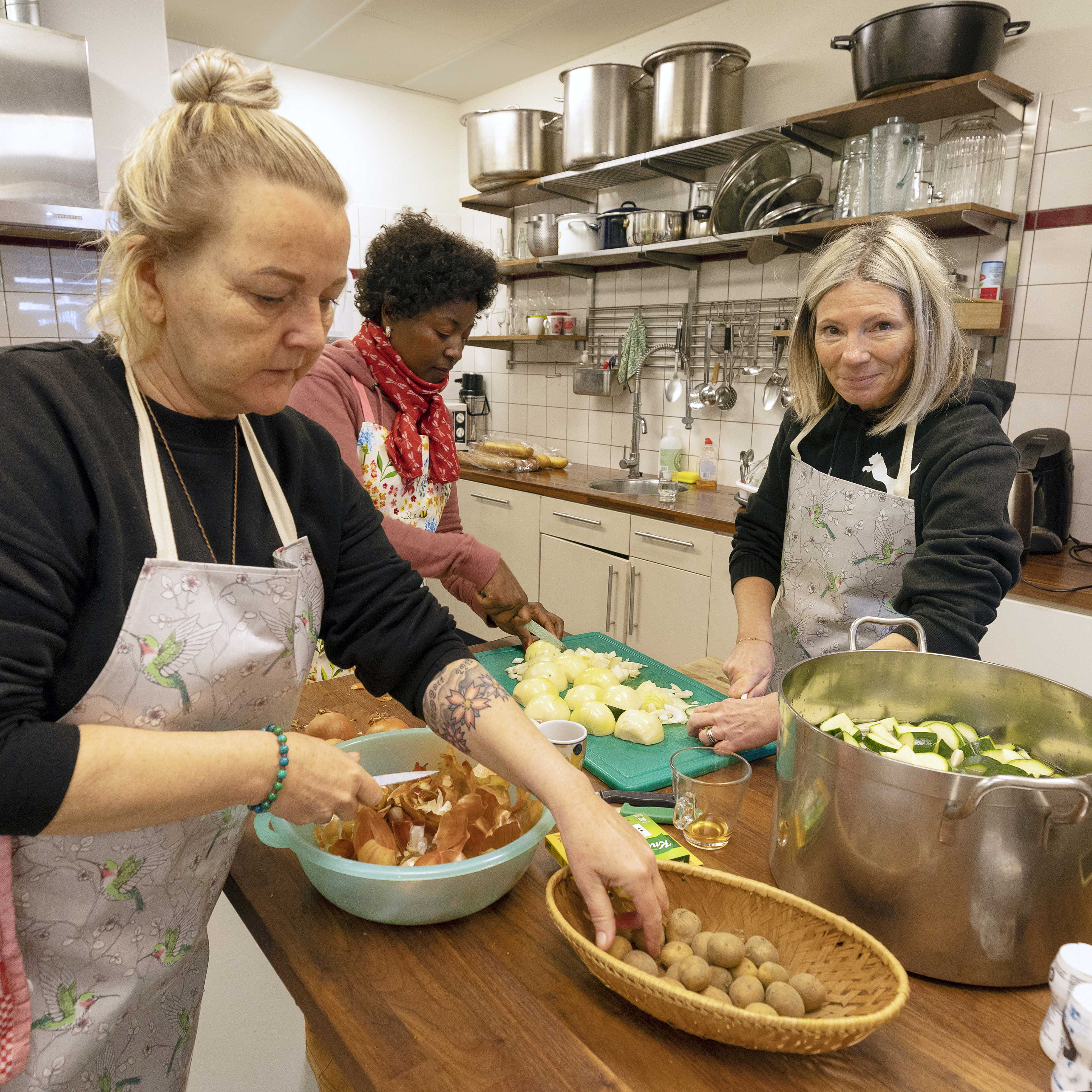 3 vrouwen dragen kookschorten. Ze snijden groente aan een keukeneiland.