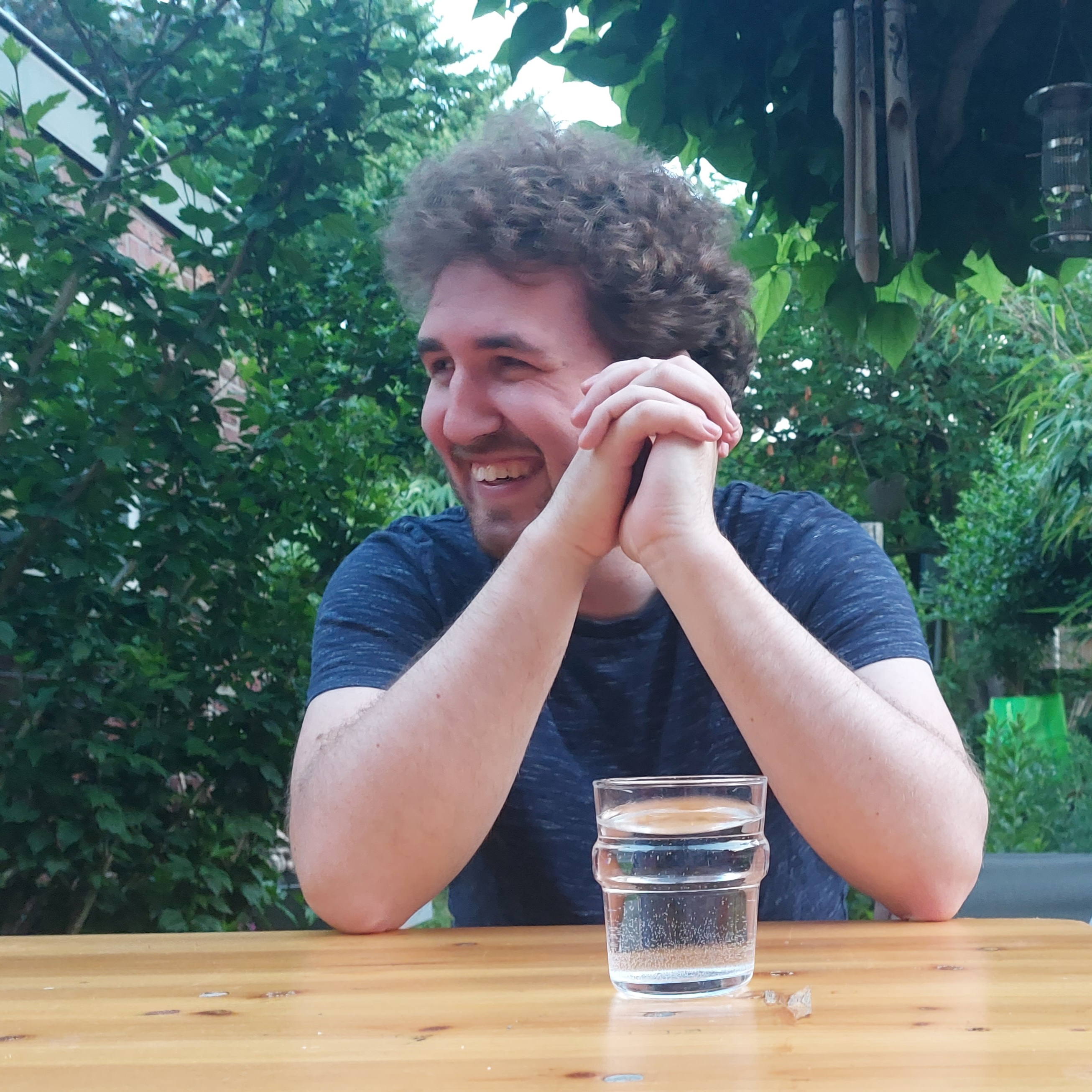 Fabian zit achter een tafel met een glas water voor zich. Hij lacht naar iemand rechts van hem. Hij heeft zijn handen voor zich gevouwen. Op de achtergrond is een tuin te zien.